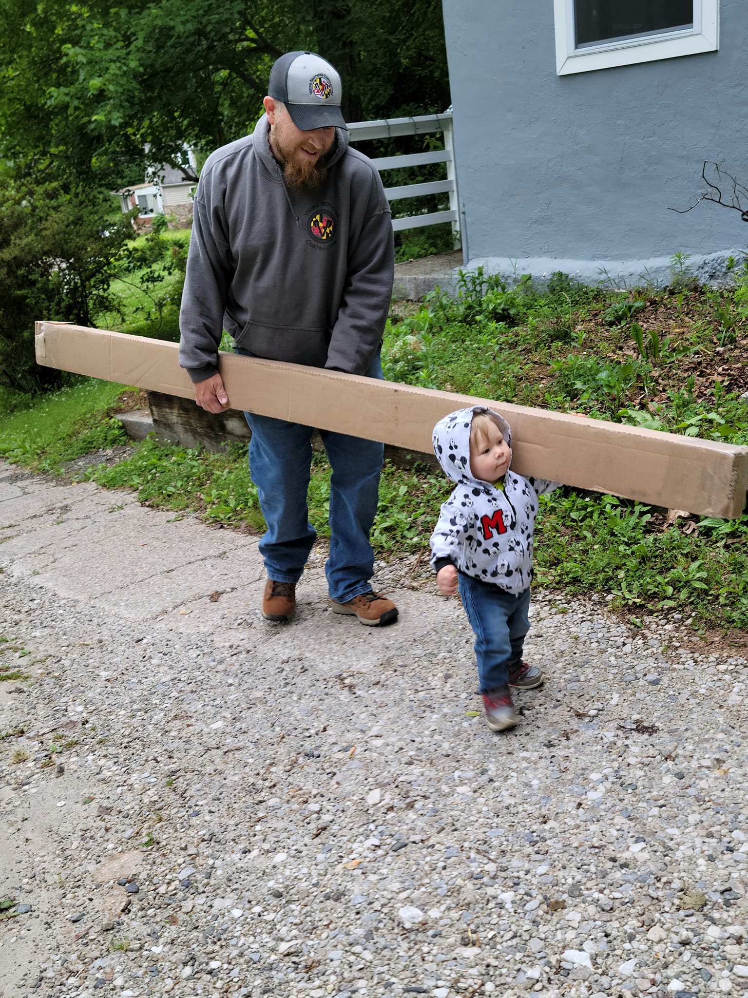 An adult and a child walk on a gravel path. The adult, wearing a cap and hoodie, carries a long, rectangular box. The child, in a hooded jacket and jeans, walks ahead, looking down.